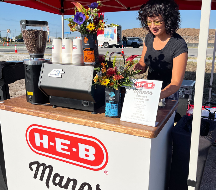 Barista behind a H-E-B custom-branded mobile coffee cart with coffee-making equipment and flowers.
