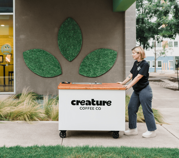 Barista at an event, pushing a 'creature coffee co' mobile coffee cart outside an office building with green leaf decorations.