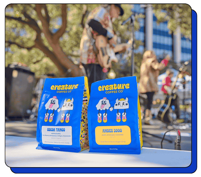 Two 'creature Coffee Co.' coffee bags on a table with a blurred outdoor concert scene in the background.