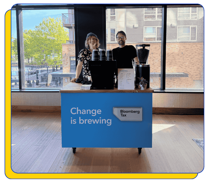 Two event baristas behind a blue counter with 'Bloomberg Tax' branding, featuring a mobile coffee cart and 'Change is brewing' text. Custom-branded coffee catering cart.