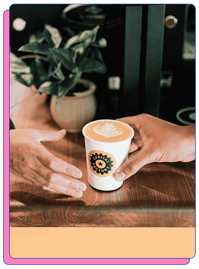 Two hands exchanging a coffee cup with latte art, and a custom-printed logo on the cup, on a wooden surface, with a plant and coffee machine in the background.