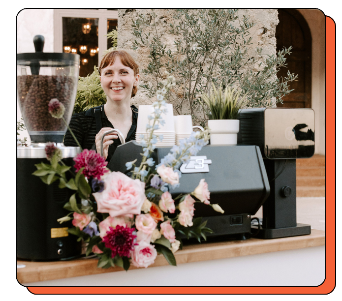 Woman barista smiling and standing behind a floral display with a coffee cart,  serving lattes at the event and plants in the background