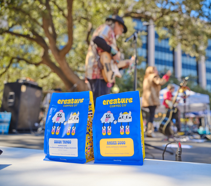 Two 'creature Coffee Co.' coffee bags on a table with a band playing in the background.