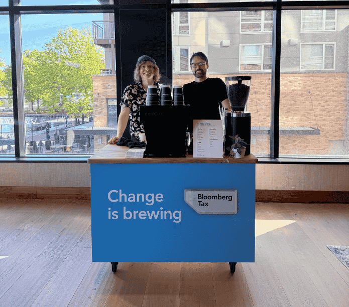Two people behind a custom mobile espresso bar with a Bloomberg Tax custom cups, menu and cart, in a modern office setting.