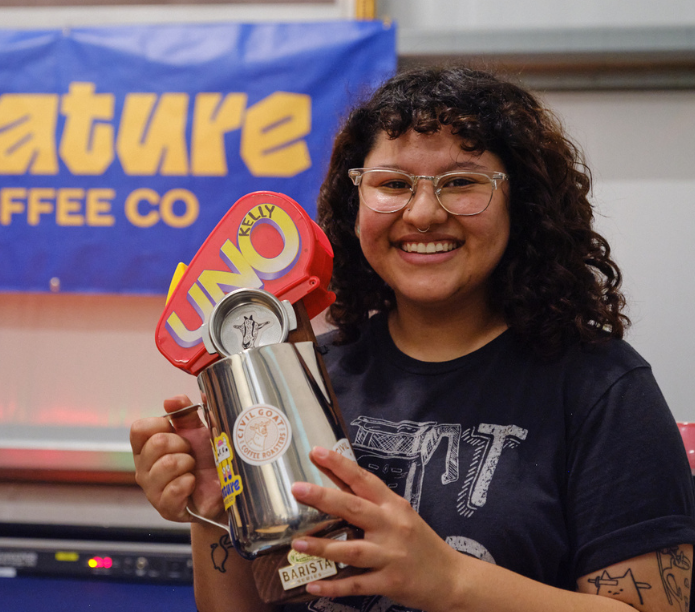 Champion Latte Art Barista holding a silver cup with colorful design in front of a 'Creature Coffee Co' banner
