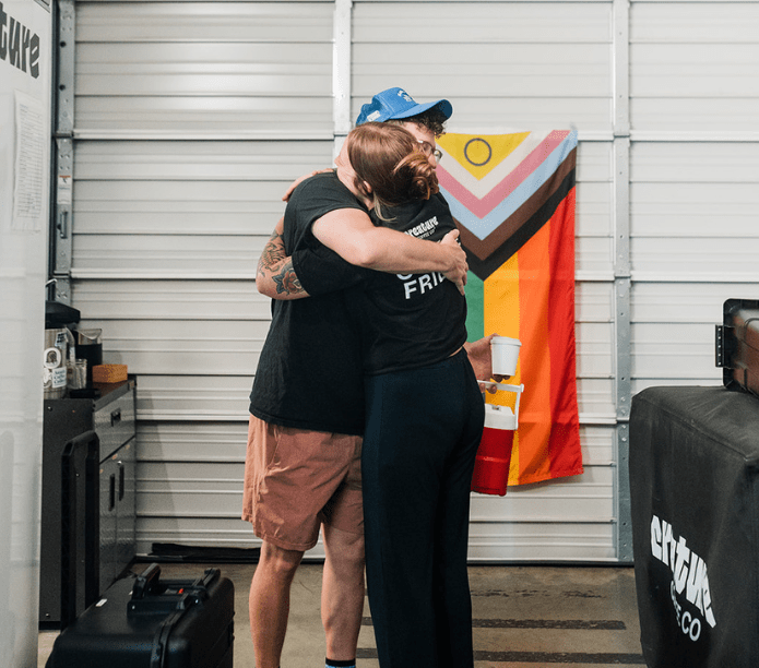 Two baristas hugging in a warehouse unit with a colorful flag in the background before working at an event with a coffee catering team