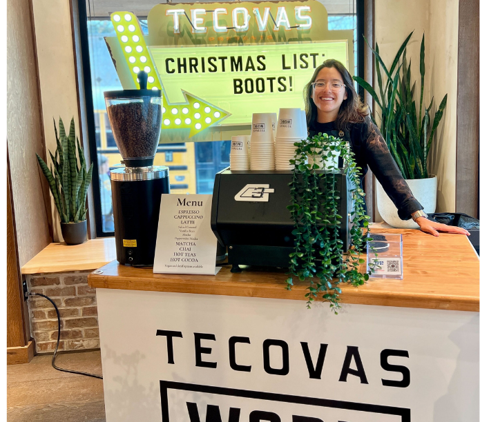 Mobile barista standing behind a mobile espresso bar with 'TECOVAS' branding, surrounded by plants and coffee catering equipment.