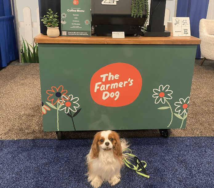 Dog standing in front of a custom-branded green mobile espresso bar with 'The Farmer's Dog' branding on the menu, cups and cart.
