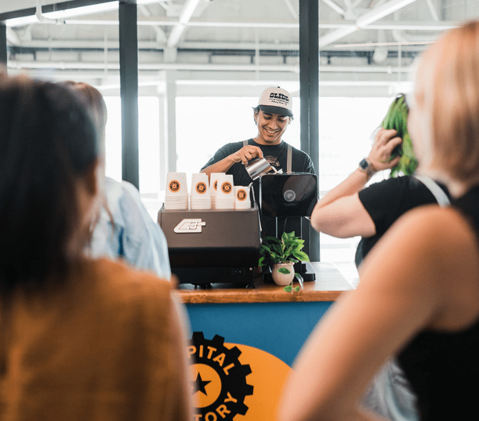Mobile barista pouring a latte drink at a counter with 'Capital City Story' branding on the menu, cups and custom coffee cart, surrounded by happy people.
