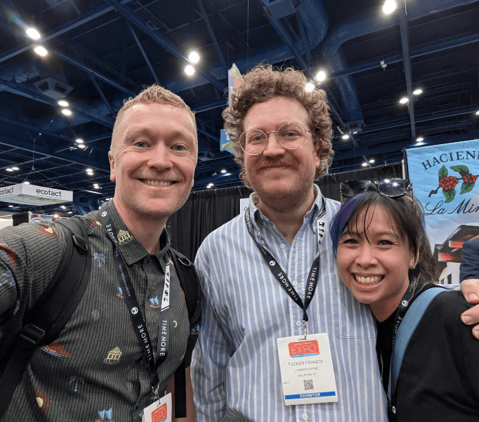 Three friends posing together at a SCA EXPO convention with a booth in the background
