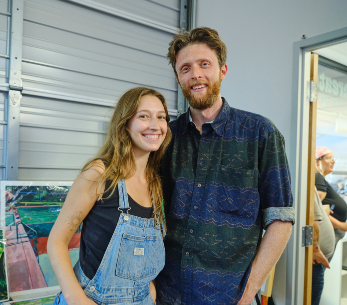 Two people standing together in a casual setting with a painting in the background