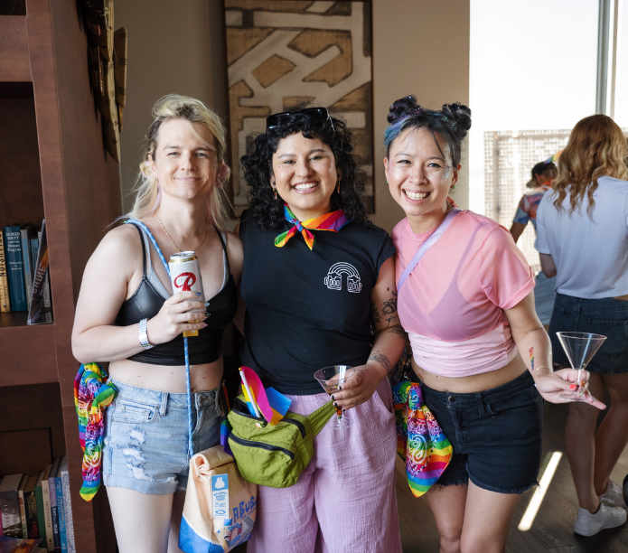 Three friends posing together in a casual setting with drinks and colorful accessories.