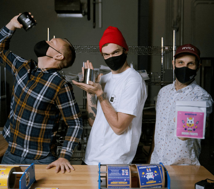 Three coffee roasters in a roasting setting, two wearing masks, one holding a pink container.