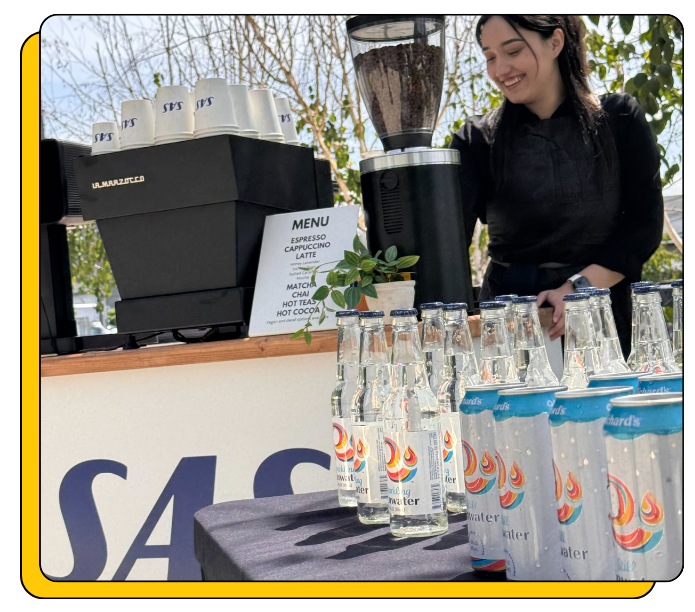 Barista an an event behind a mobile espresso bar and coffee cart, with a water station with branded cups, cart and bottles.