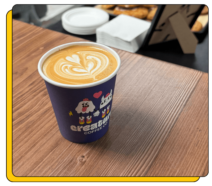 Compostable coffee cup with latte art on a wooden table, featuring the 'Creature Coffee' brand at an event. 