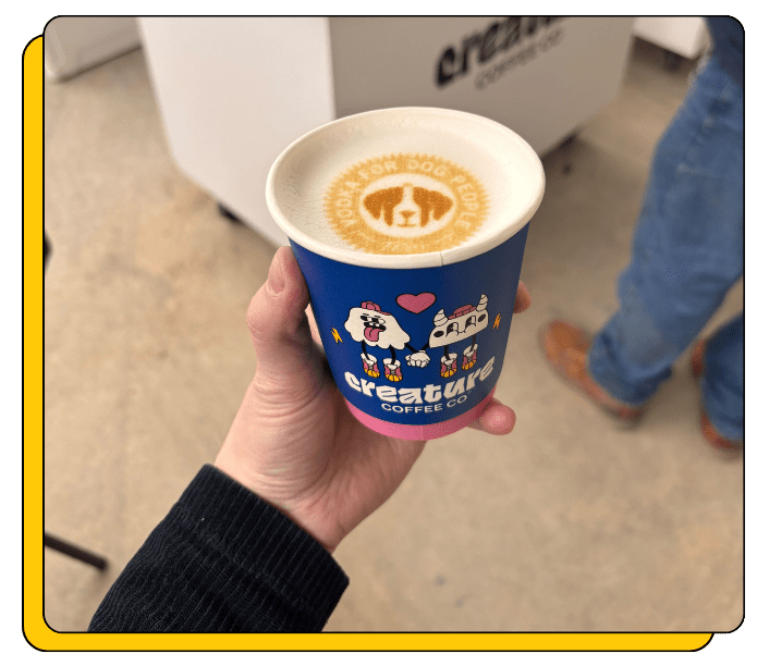 Hand holding a blue 'creature Coffee Co.' cup with a custom design printed on the latte foam of a cappuccino at a coffee catering event.