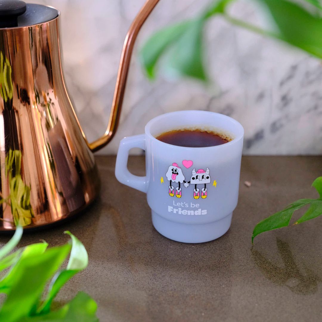 White mug with cartoon characters and 'Let's be Friends' text next to a bronze kettle on a marble surface. GIft from Specialty Coffee Roaster.
