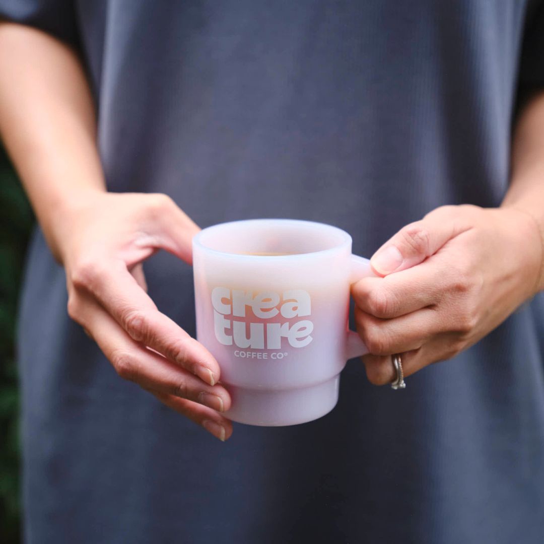 Person holding a 'Crea ture Coffee Co.' mug with steam, blurred background. GIft from Specialty Coffee Roaster.