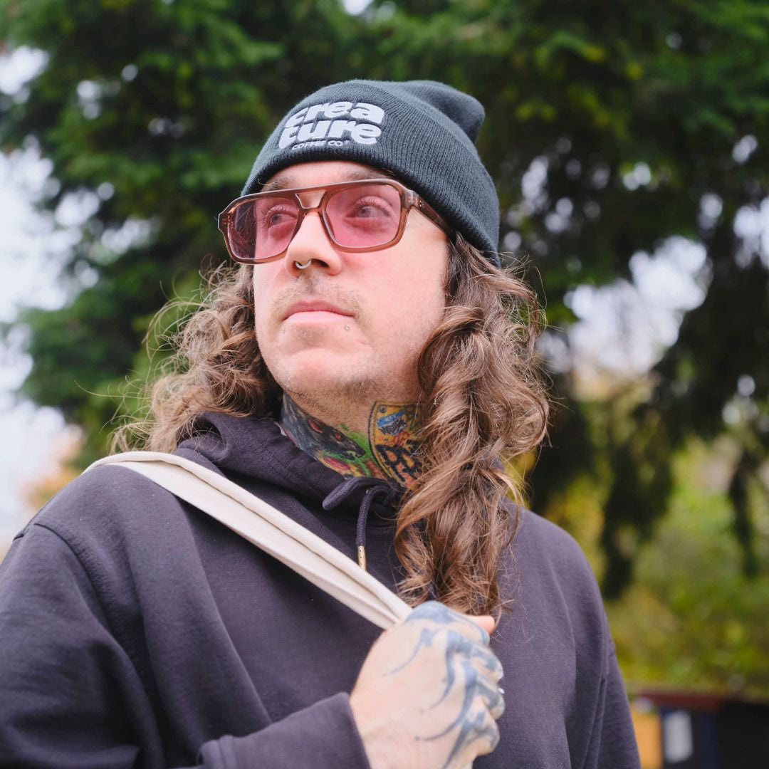 Person with long hair and tattoos wearing a beanie and sunglasses outdoors. GIft from Specialty Coffee Roaster.