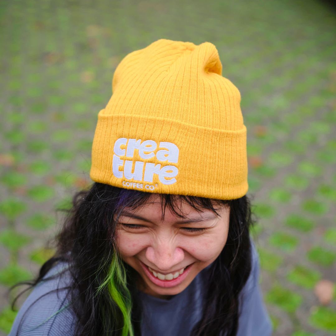 Person wearing a yellow beanie with 'creature coffee' branding outdoors. GIft from Specialty Coffee Roaster.