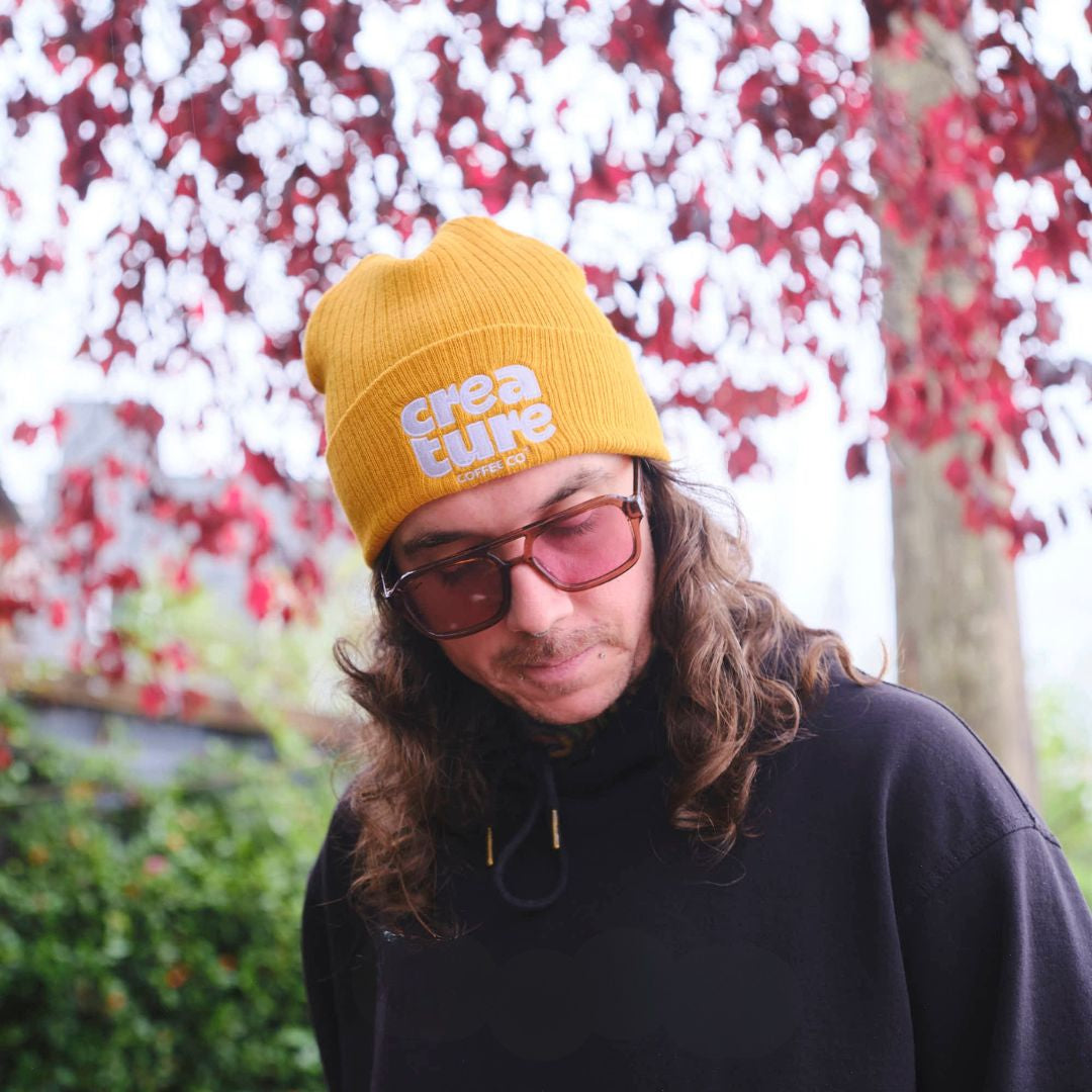 Person wearing a yellow 'creature coffee' beanie with a blurred floral background