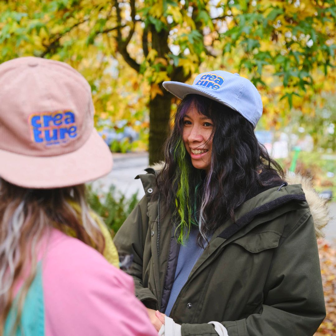 Two people wearing hats with 'creature coffee' branding, standing outdoors with trees in the background.