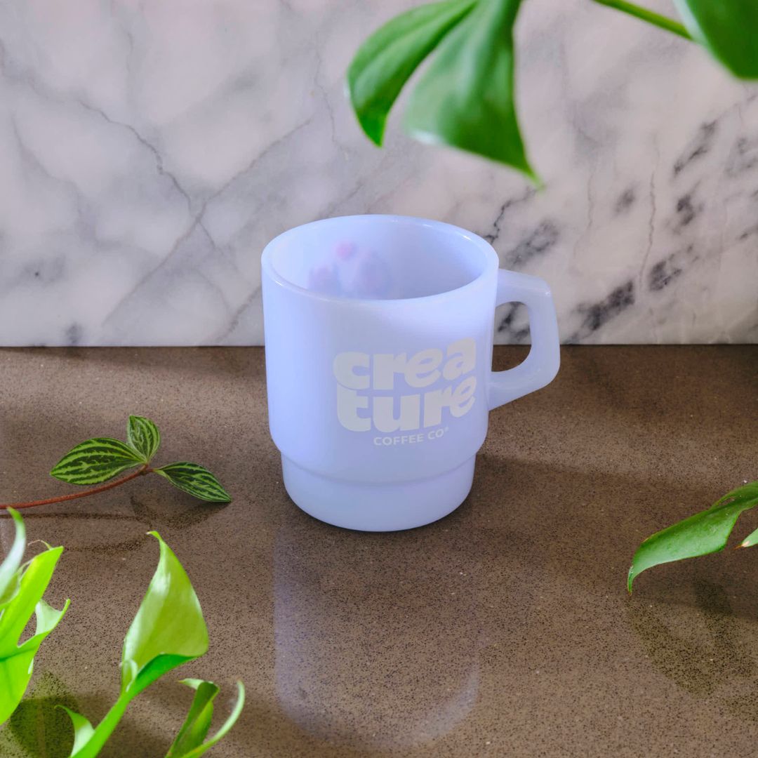White frosted mug with 'creature coffee co.' text on a marble surface with green leaves. GIft from Specialty Coffee Roaster.