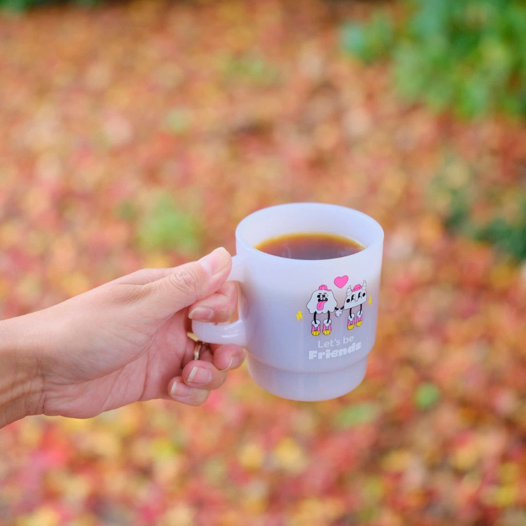 Hand holding a white mug with cartoon cows and text against a blurred natural background. GIft from Specialty Coffee Roaster.