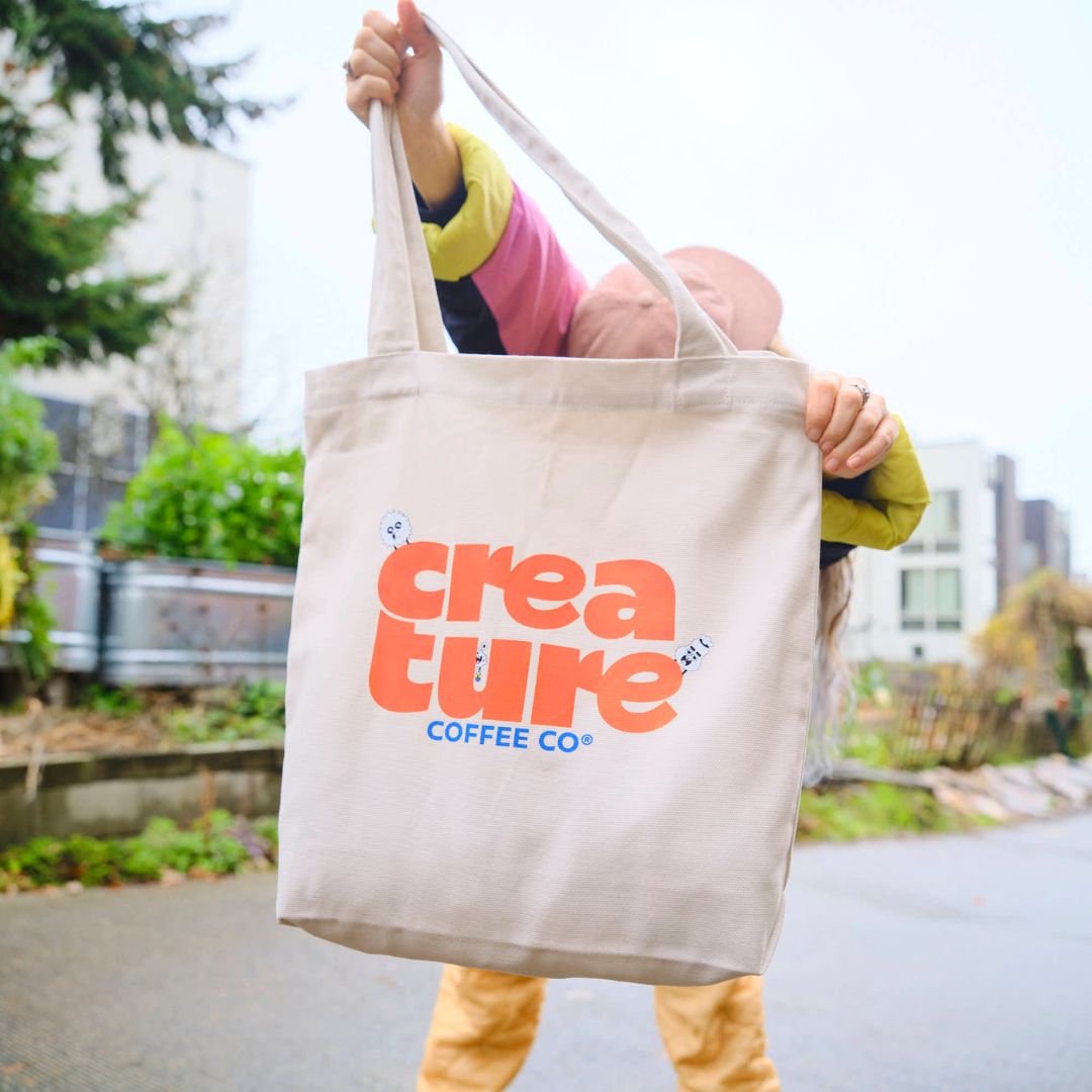 Person holding a tote bag with 'creature COFFEE CO' logo outdoors. GIft from Specialty Coffee Roaster.