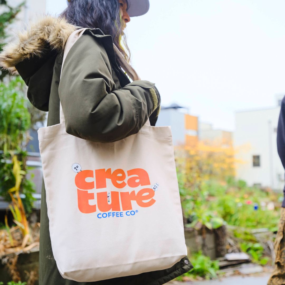 Person holding a tote bag with 'creature coffee co' branding outdoors. GIft from Specialty Coffee Roaster.