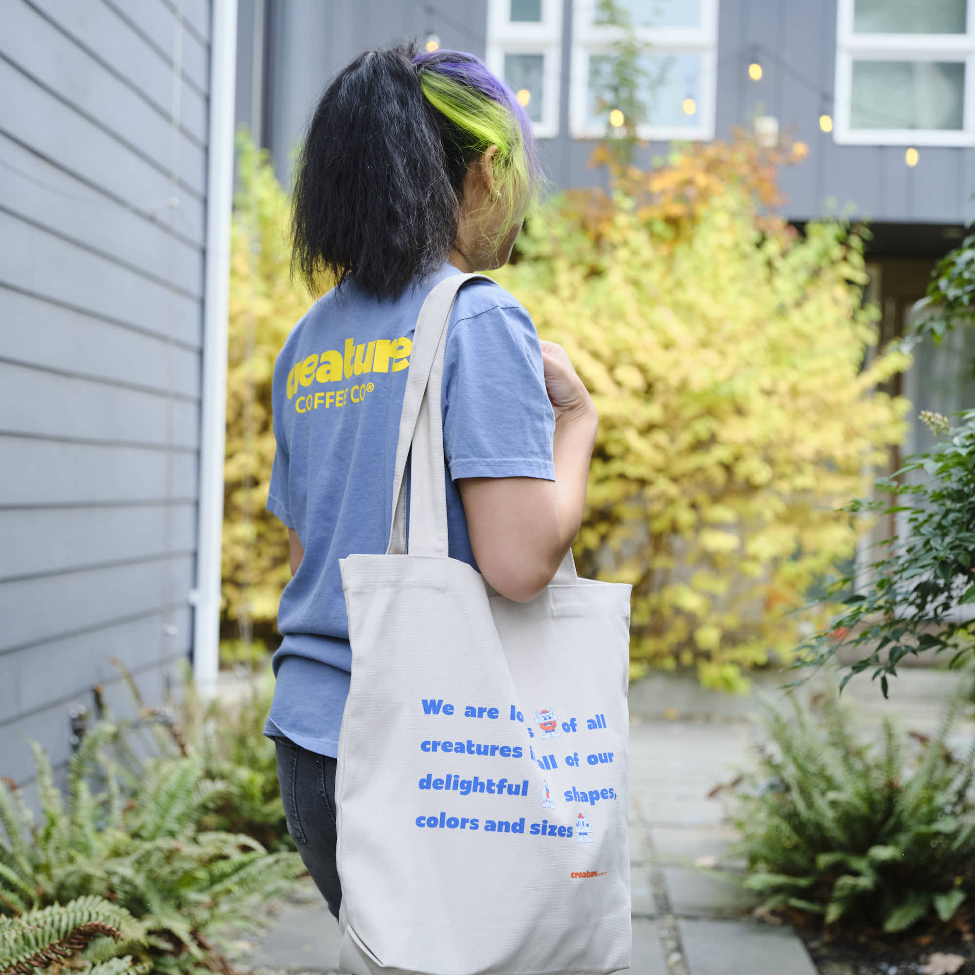 Person holding a tote bag with creature coffee text, standing outdoors near a building.
