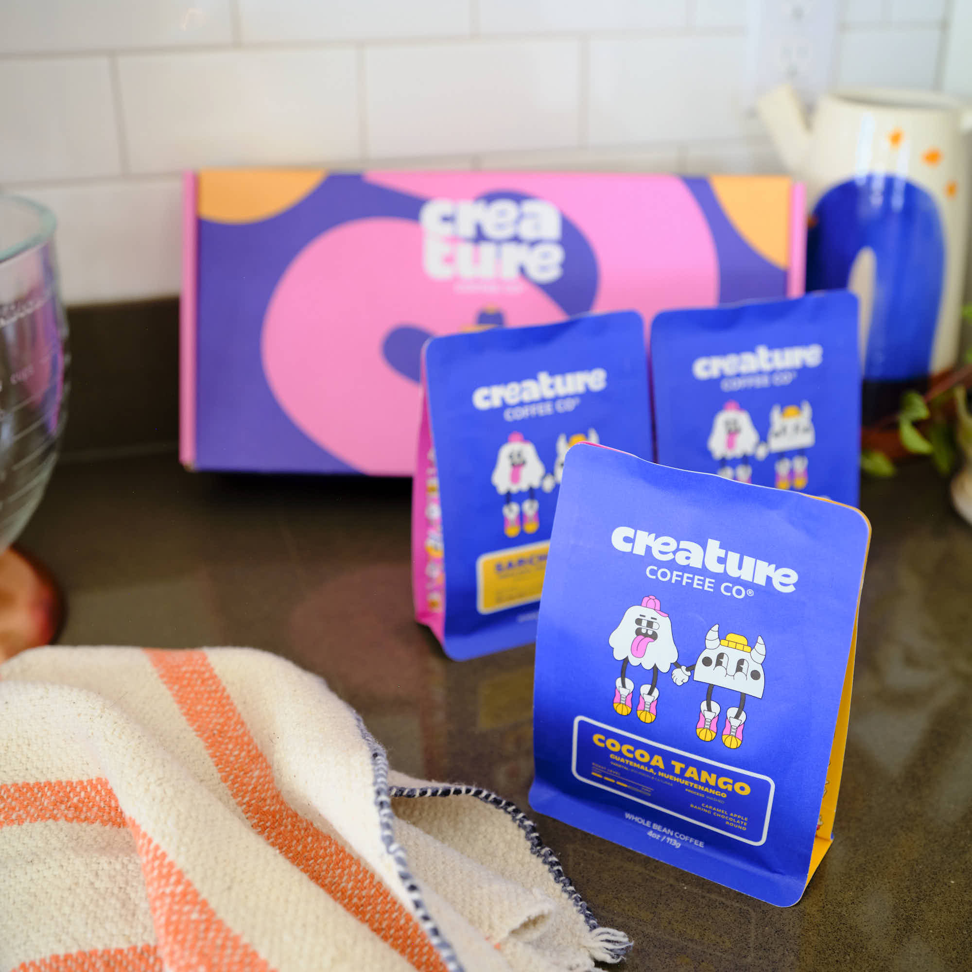 Blue 'creature Coffee Co.' packages on a kitchen counter with a colorful box in the background. Gift box for coffee lovers. 