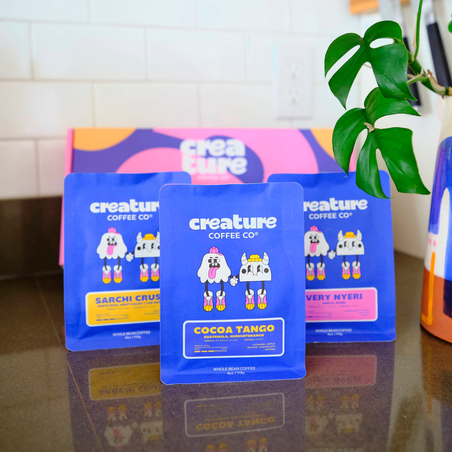 Three blue creature coffee bags with cartoon characters on a kitchen counter with a plant and colorful box in the background. Gift box for coffee bags. 