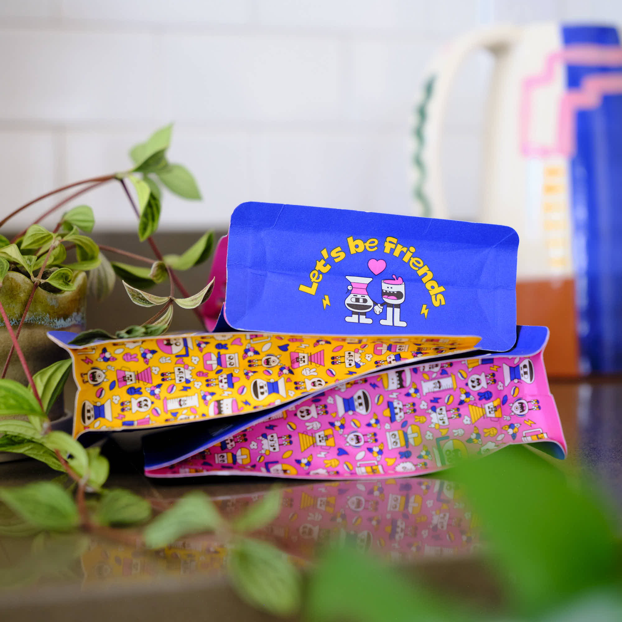 Colorful creature coffee pouches with cartoon designs on a surface with plants in the background