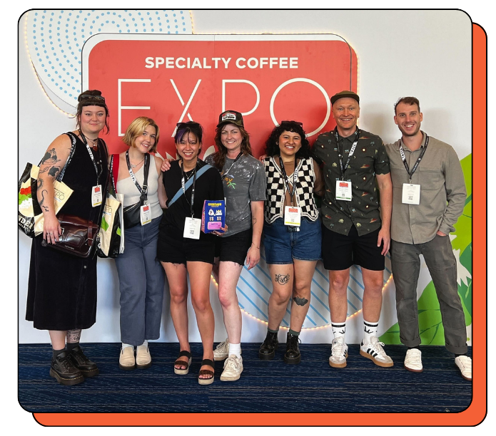 Group of baristas and coffee roasters posing in front of a Specialty Coffee Expo backdrop. Team Creature Coffee are very happy and smiling. 